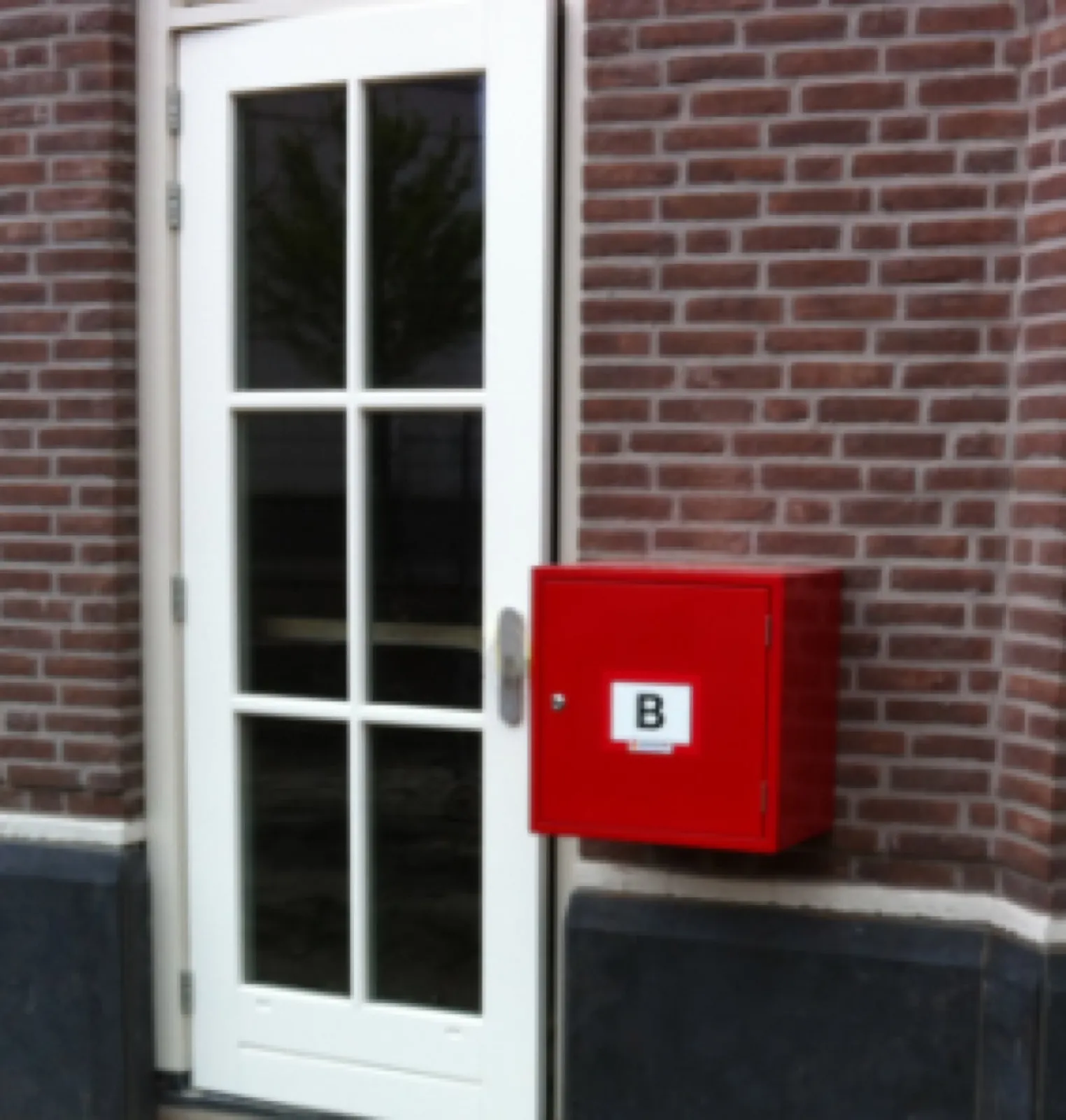 Rode kast met B-pictogram naast een voordeur in een bakstenen gevel